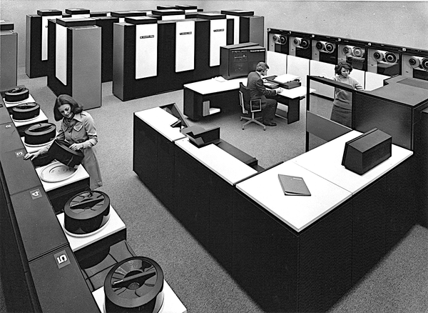 A picture showing a man seated at the control desk of a Multics installation in a large room